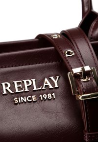 Sac en simili cuir bordeaux avec un logo doré "REPLAY" et "SINCE 1981." Dispose d'une forme structurée, d'une bandoulière accentuée et de quincaillerie métallique.