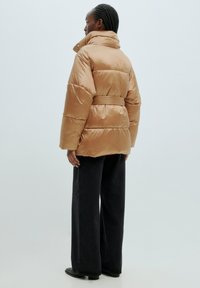 Gepolsterte, mit Gürtel versehene Jacke aus hellbraunem Satinmaterial. Verfügt über einen hohen Kragen und eine übergroße Silhouette. Getragen mit weiten schwarzen Hosen.