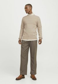 Pull en tricot beige avec des motifs côtelés horizontaux, superposé sur une chemise rayée blanche et beige, associé à un pantalon gris et des bottes marron.