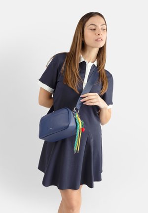 Donna con abito blu navy che indossa una borsa a tracolla blu navy con nappe colorate, guardando leggermente verso destra su uno sfondo semplice.