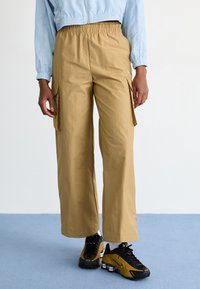 Smėliniai plačiagyviai kroviniai su elastine juosmeniu, turintys šonines kišenes ir glotnią tekstūrą, derinami su juodais sportiniais batais.