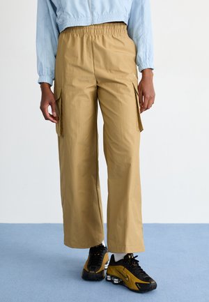 Pantaloni cargo beige a gamba larga con vita elastica, dotati di tasche laterali e una texture liscia, abbinati a scarpe sportive nere.