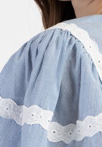Primo piano di una blusa a righe blu e bianche con maniche a sbuffo, caratterizzata da un delicato bordo in pizzo bianco lungo la cucitura della spalla.