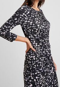 Robe noire ornée d'un motif de formes abstraites blanches et grises, à manches longues, avec une silhouette ajustée et des côtés froncés.