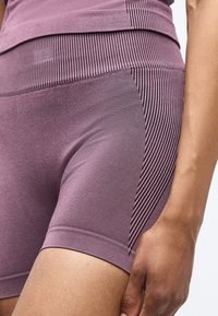Κοντινή λήψη ατόμου που φοράει εφαρμοστά βιολετί σορτς με ραβδωτή υφή και ριγέ πλευρικές λωρίδες, το χέρι ανάπαυεται στο μηρό.