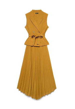 Gilet senza maniche giallo senape con cintura e risvolti abbinato a una gonna lunga plissettata a vita alta dello stesso colore su sfondo bianco.