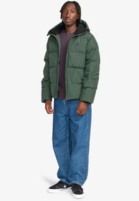 Giubbotto imbottito in nylon verde scuro con zip, fodera in pile nera e cappuccio, abbinato a jeans blu a taglio ampio e sneakers nere.