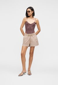 Γυναίκα που στέκεται με τα χέρια πίσω από την πλάτη, φορά γυαλιά ηλίου, μοβ φόρεμα με κουμπιά, μπεζ σορτς και μαύρα σανδάλια με τακούνι σε λευκό φόντο.