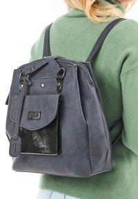 Une femme portant un pull vert clair porte un grand sac �à dos texturé gris avec une poche avant noire brillante et des détails en cuir.