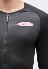 Gros plan d'une personne portant un maillot de cyclisme noir texturé avec une fermeture éclair à l'avant et le logo rose "Isadore" sur la poitrine.