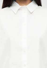 Γυναίκα με λευκό πουκάμισο με κουμπιά και μυτερό γιακά, εμφανίζεται από το λαιμό έως το άνω μέρος του κορμού.