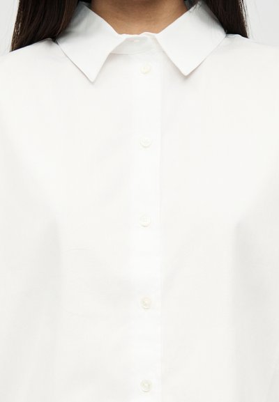 Kvinne iført hvit skjorte med knapper og spiss krage, vist fra nakke til overkropp.