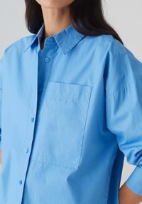 Camicia azzurro chiaro realizzata in tessuto liscio, con chiusura a bottoni sul davanti, colletto a punta e una tasca sul petto. Maniche corte e morbide.