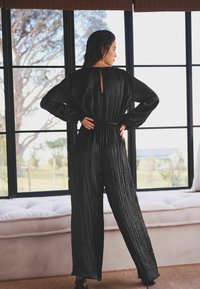 Schwarzer plissierter Jumpsuit mit langen Ärmeln, tailliertem Bund und Schlüsselloch-Rücken. Der Stoff hat eine glatte Textur. Das Modell posiert mit den Händen in den Hüften.