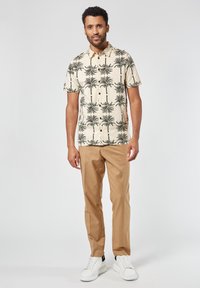 Korte mouwen beige shirt met groen palmbomenpatroon, gecombineerd met tan broek en witte sneakers. Eenvoudig button-down ontwerp.