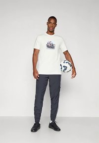 Hvid T-shirt med grafisk tryk, kombineret med mørkegrå træningsbukser med hvide striber i siden og sorte sportssko. Holder en fodbold.