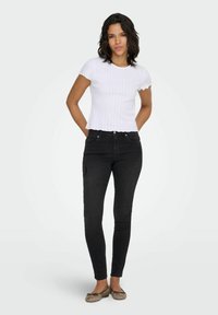 Valge soonikkoega lühikeste varrukatega topp laineliste servadega, kombineeritud mustade kitsaste teksade ja pruunide tasapõhjadega helehalli tausta vastu.