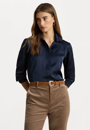 Blusa di raso blu navy con design a bottoni, maniche lunghe arrotolate fino al gomito, abbinata a pantaloni in velluto a coste marrone chiaro e una cintura color sabbia.