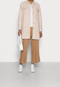 Giacca oversize beige pelosa con due tasche frontali, camicia bianca, pantaloni larghi color beige e borsa bianca. Le sneakers completano il look.