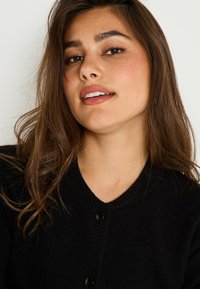 Cardigan noir avec un col en V, présentant un tissu texturé, un col côtelé et des boutons noirs brillants. Les cheveux sont ondulés, encadrant partiellement le visage.