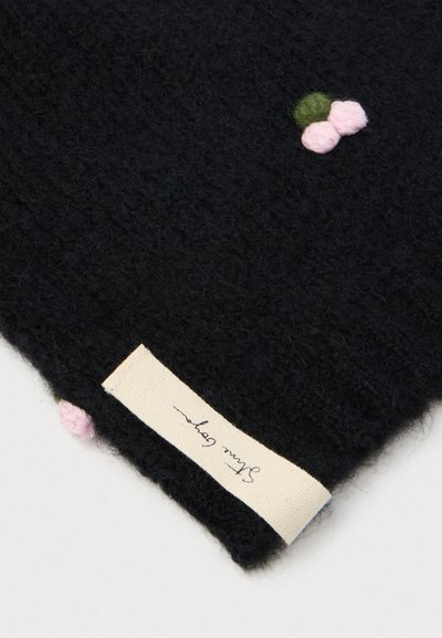 Cachecol de lã preta tricotado com duas flores bordadas em rosa e verde e um rótulo cosido com texto manuscrito. Textura suave, forma retangular.