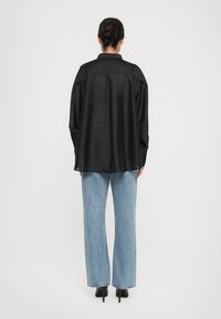 Kvinne med hår i bun, iført en løs svart skjorte med knapper, lyseblå rette jeans og svarte høyhælte sko, stående med ryggen til.