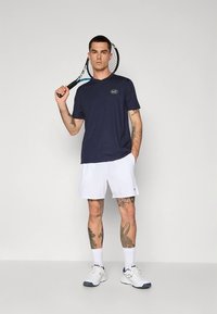 Mørkeblå sports t-skjorte med korte ermer og logo, kombinert med hvite shorts og hvite treningssko, holder en tennisracket.