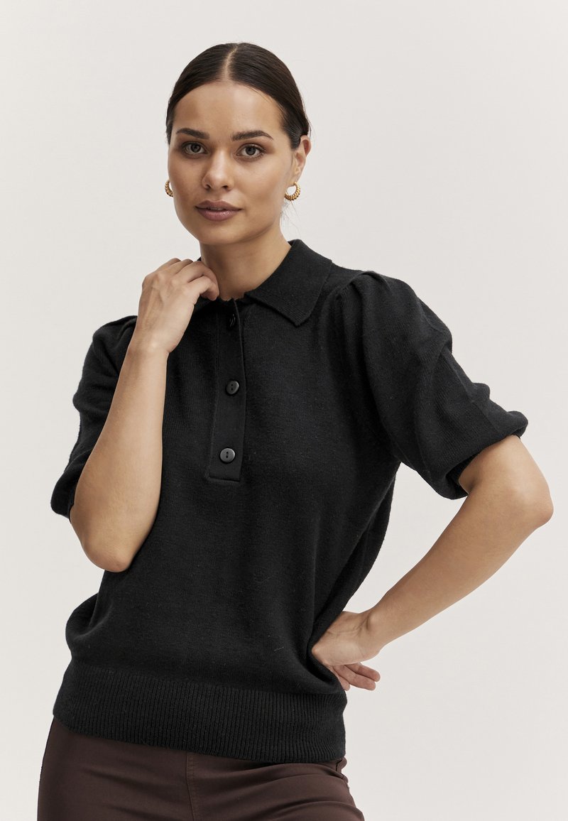 Maglione polo nero a maniche corte con colletto, pattina a tre bottoni e orlo a coste. Realizzato in un tessuto a maglia testurizzato.