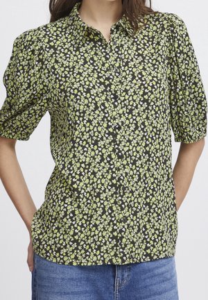 Kobieta ubraną w czarną bluzkę na guziki z małym zielono-białym kwiatowym wzorem i bufkami na krótkich rękawach, do tego niebieskie dżinsy.