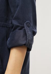 Manche bleu marine retroussée maintenue par une patte boutonnée sur le bras d'une personne, sur un fond clair uni.