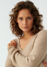 Beige pletený svetr s výstřihem do V a žebrovanými manžetami, který má měkkou texturu a volný střih. Model pózuje se zkříženýma rukama.