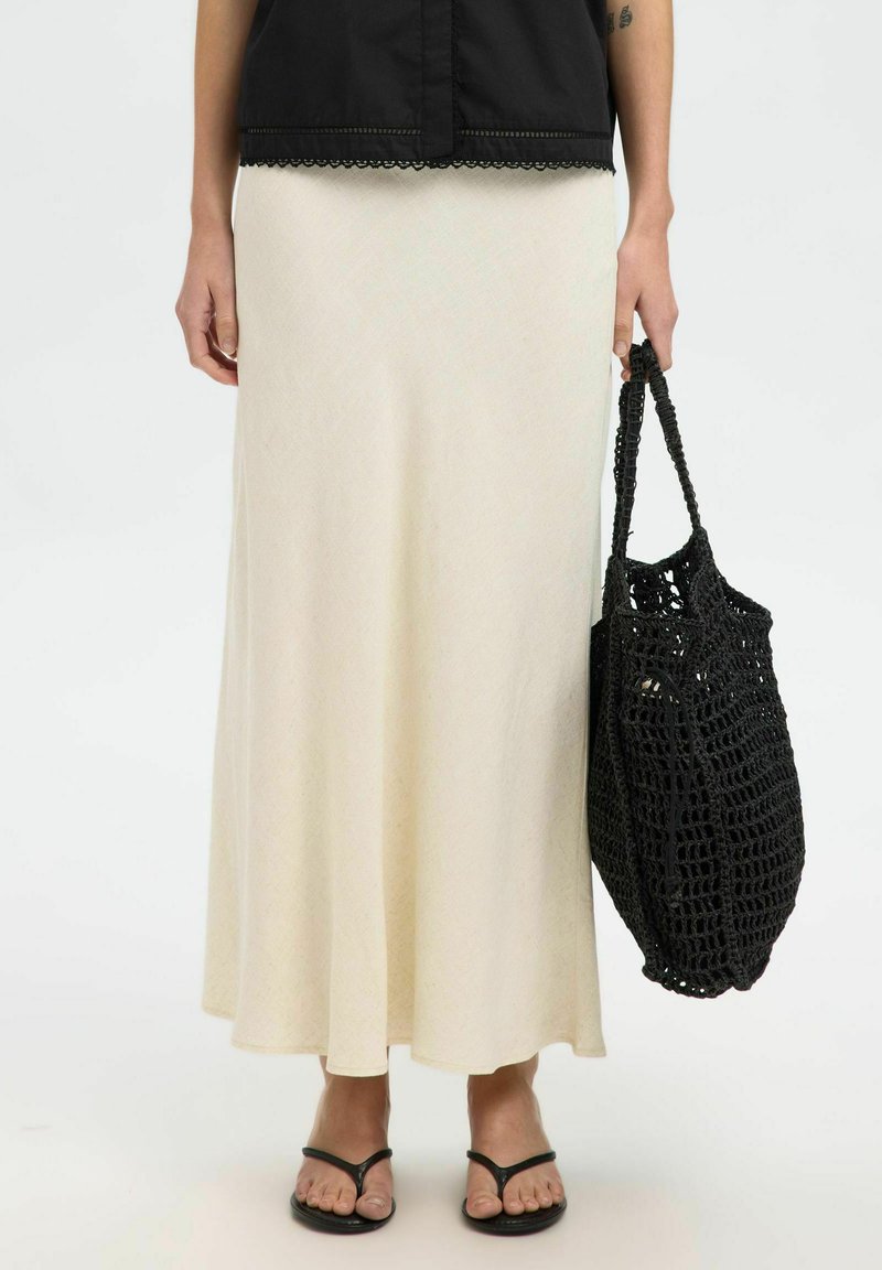Jupe maxi couleur crème avec une texture lisse, associée à un sac fourre-tout en crochet noir et des tongs noires.