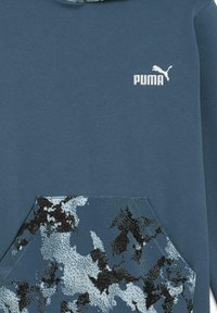 Mikina s kapucňou v námorníckej modrej farbe s vreckom s maskáčovým vzorom v odtieňoch modrej a čiernej. Na hrudi sa nachádza biele logo Puma.
