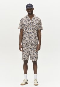 Short-sleeve collared shirt and matching shorts in beige with a blue bird pattern, paired with a navy cap and sneakers.