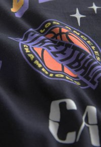 Svart tyg visar en basketboll-logotyp med lila och orange detaljer. Inkluderar stjärnor och texten "Los Angeles" och "Champions."