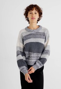 Maglione a righe grigio e bianco con scollo rotondo, texture voluminoso e polsini a coste, indossato con un pantalone nero uniforme.