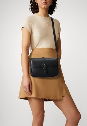 Femme portant un haut beige côtelé à manches courtes et une jupe portefeuille couleur camel, portant un sac bandoulière en cuir noir avec une fermeture à boucle.
