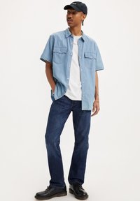 Camisa de mezclilla de manga corta azul claro con dos bolsillos en el pecho, llevada sobre una camiseta blanca. Jeans de pierna recta azul oscuro y zapatos negros.