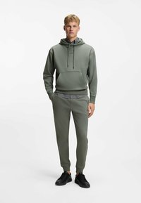 Green hoodie with drawstrings and front pocket, paired with matching tapered pants. Black sneakers complete the look. Casual, smooth fabric.