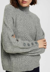 Pull en tricot gris avec col montant, corps texturé et gros boutons ronds gris aux poignets. Fil doux et épais avec des détails côtelés.