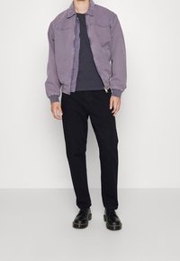 Fialová bombardérka s předním zipem, žebrovanými manžetami a límcem, spárována s černými kalhotami a černými botami. Hladká textura, volný střih.