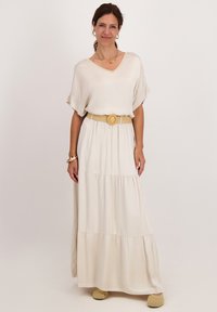 Crème kleurige, gelaagde maxi-jurk met een V-hals en korte mouwen, getailleerd met een geweven riem. Model draagt gele instap schoenen.