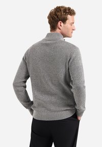 Grijze gebreide trui met een hoge kraag, voorzien van een gestructureerd patroon en geribbelde mouwen, gedragen over zwarte broeken.