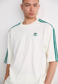 T-shirt alb din bumbac cu accente verzi, cu trei dungi pe umeri și un mic logo Adidas verde pe piept. Croială lejeră, gât rotund.
