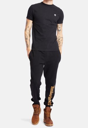 Mann trägt schwarze Timberland-Sweatpants mit gelbem Logo, schwarzes enganliegendes T-Shirt, braune Lederstiefel und sichtbare Arm-Tattoos, steht.