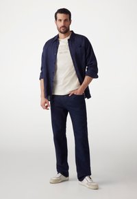 Camisa de botones azul marino sobre una camiseta gráfica de color crema claro, combinada con jeans azul oscuro y zapatillas blancas. Diseño sencillo, ajuste relajado.