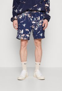 Marineblå shorts med et fargerikt fuglemønster, med elastisk midje, sidelommer og en avslappet passform. Brukt med hvite sneakers og lyse sokker.