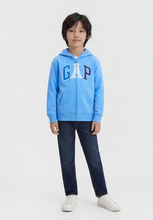 Dieťa stojí čelom dopredu, oblečené v modrej mikine GAP s kapucňou, tmavých džínsoch a bielych teniskách na jednoduchom svetlom pozadí.