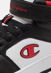 Czarno-białe trampki z teksturowanymi skórzanymi cholewkami, czerwonymi akcentami, logo z boku oraz sznurowanym zapięciem z rzepem.