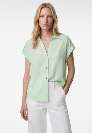 Kobieta o blond włosach, ubrana w jasnobeżową koszulę na krótki rękaw z guzikami i białe spodnie, stojąca z jedną ręką w kieszeni.